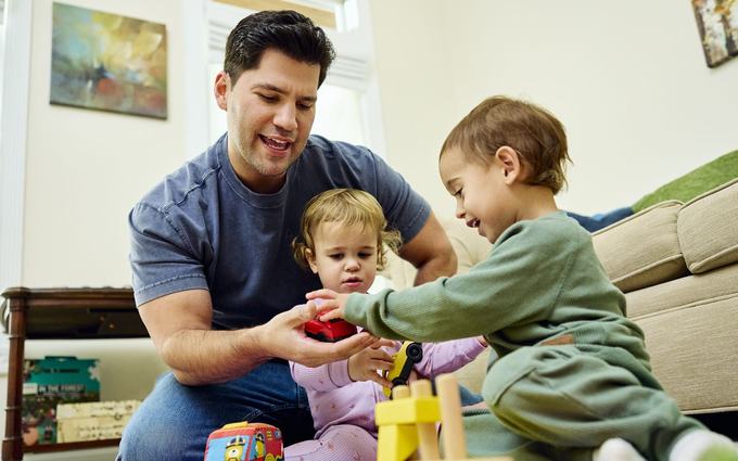 Un padre juega en el suelo de la sala con dos niños pequeños, entre bloques de madera y carritos de juguete.