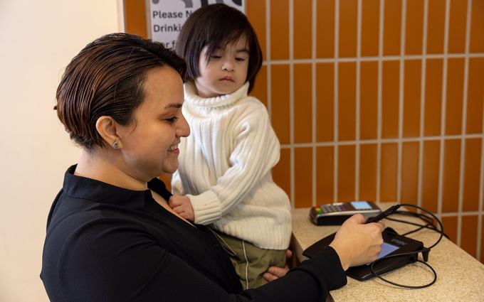 Una madre con su hijo pequeño en brazos firma en una tableta electrónica sobre el mostrador de una clínica.