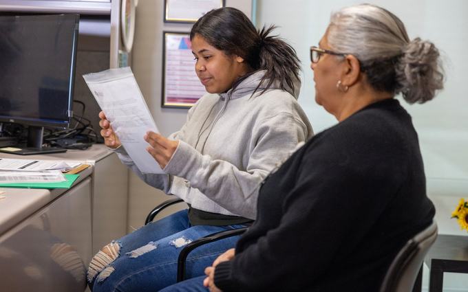 Una joven con sudadera gris lee una carta en una oficina de la clínica, sentada junto a una mujer mayor de cabello canoso.