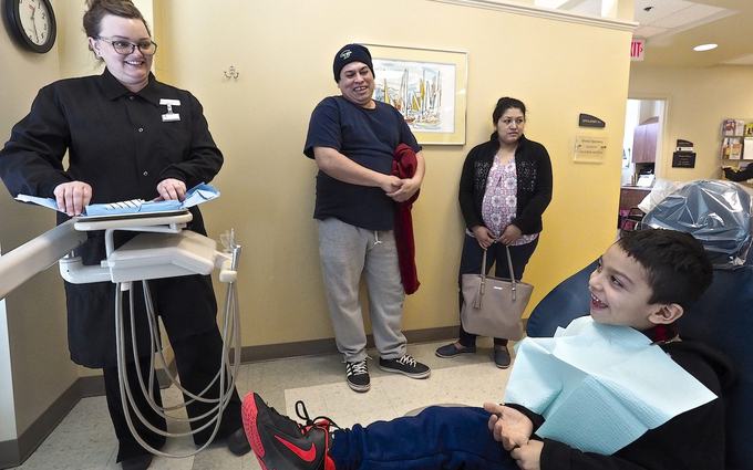 Un niño con babero sonríe en el sillón dental; una higienista prepara la bandeja y un hombre y una mujer lo miran desde la pared.