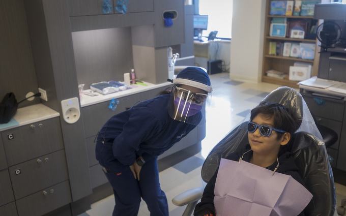 Un niño con lentes de protección azules y un babero morado sonríe en un sillón dental, junto a una higienista con careta y mascarilla.