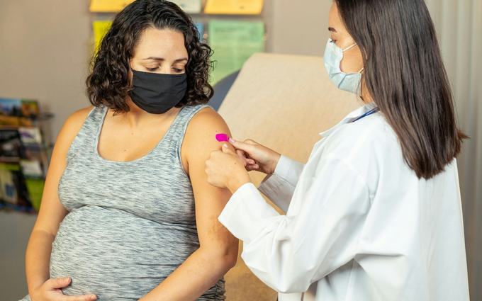 Una mujer embarazada con mascarilla mira cómo una trabajadora de salud le pone una curita rosada en el brazo, en un consultorio.