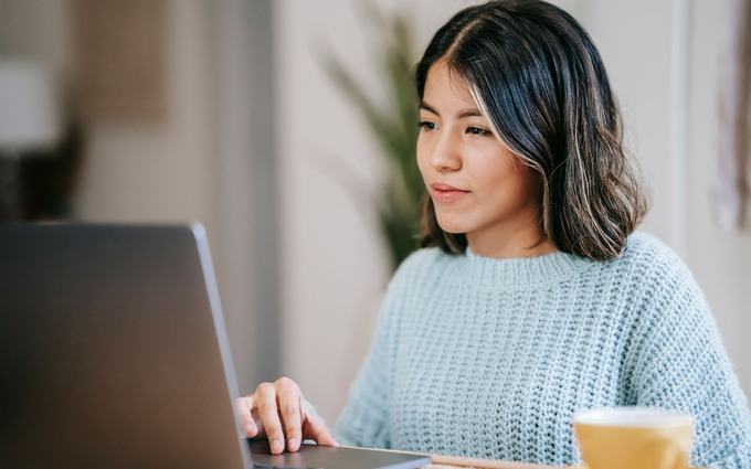 Una joven con suéter celeste escribe en una computadora portátil sobre una mesa, junto a una taza amarilla.