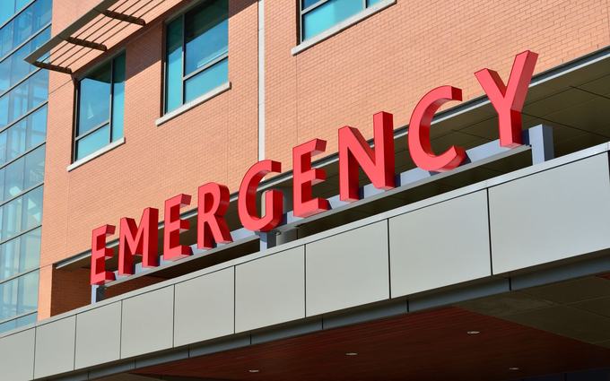 Letrero rojo que dice "Emergency" (emergencias) sobre la entrada de un hospital de ladrillo, en un día soleado.