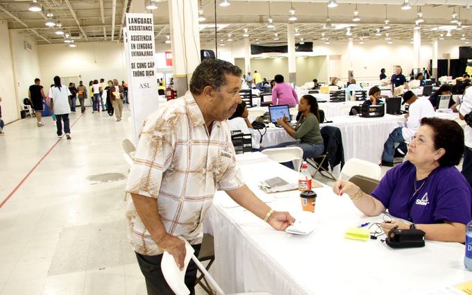 Un hombre mayor de pie habla con una intérprete sentada a una mesa; detrás, un letrero dice Language Assistance y Asistencia en lenguas extranjeras.