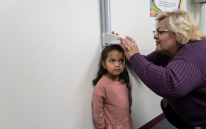 Una trabajadora de la clínica mide la estatura de una niña con vestido rosado contra una pared.
