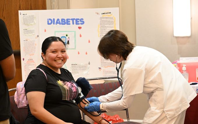 Una joven sonríe mientras una trabajadora de salud le toma la presión en un evento comunitario, frente a un cartel sobre la diabetes.