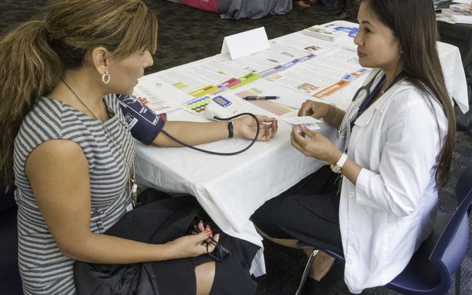 Una mujer con el brazalete de la presión en el brazo habla con una enfermera de bata blanca en una mesa con folletos de salud.