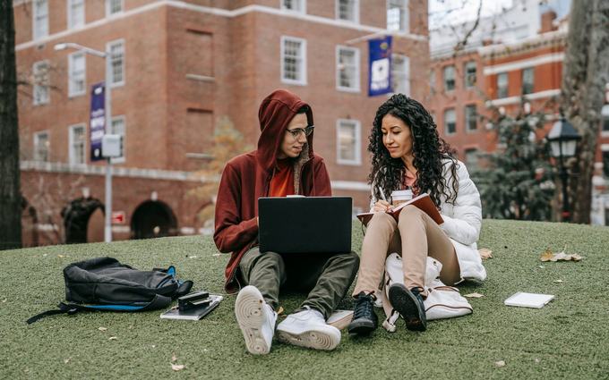 Dos jóvenes estudian en el césped de un campus, él con una computadora portátil y ella con un cuaderno, frente a edificios de ladrillo.
