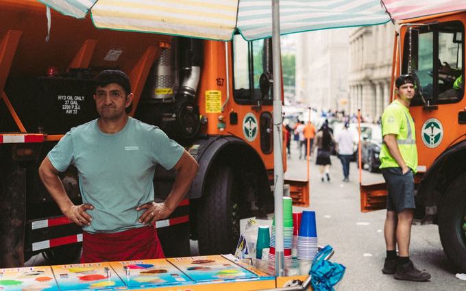 Un vendedor ambulante con camiseta verde agua y delantal rojo espera detrás de su carrito de helados en una calle de Nueva York.