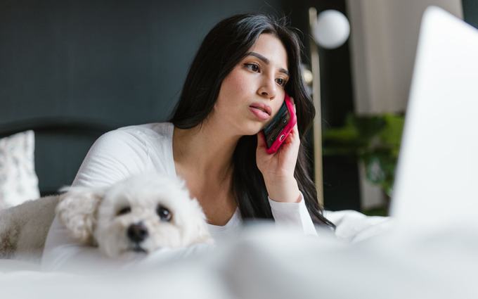 Una mujer recostada en la cama escucha con atención una llamada por teléfono, junto a un perro pequeño de pelo blanco.