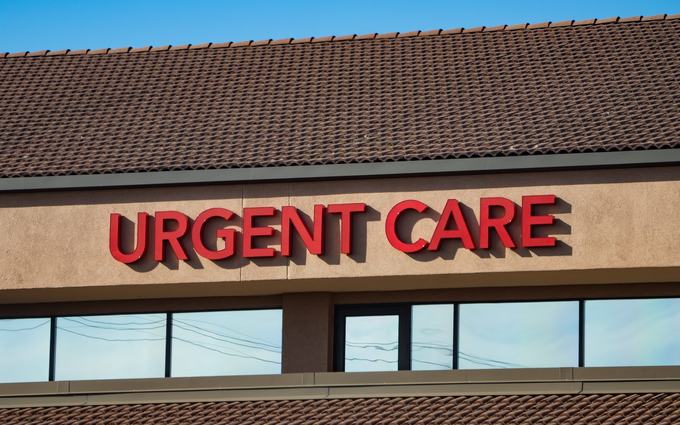 Letrero rojo que dice "Urgent Care" (atención de urgencia) en la fachada de un edificio con techo de tejas, bajo un cielo azul.