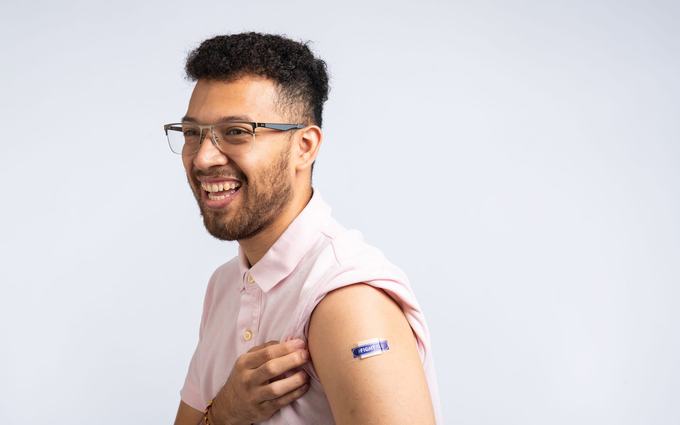 Un joven con lentes sonríe y se toca la camisa junto a una curita azul en el brazo, recién vacunado, ante un fondo gris claro.