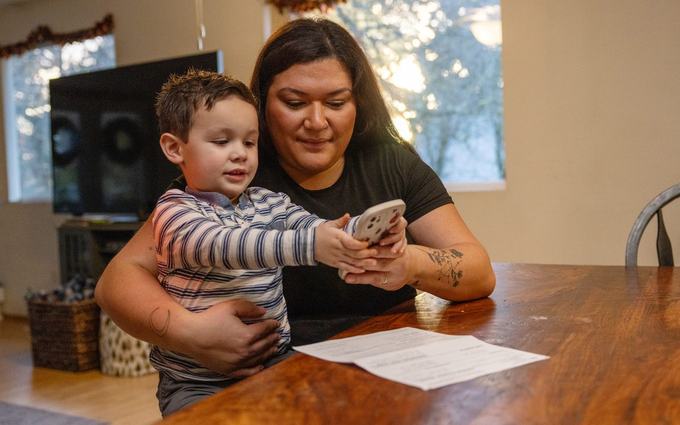 Una madre sentada a la mesa del comedor sostiene a su hijo pequeño, que agarra un teléfono; frente a ellos hay unas hojas de papel.