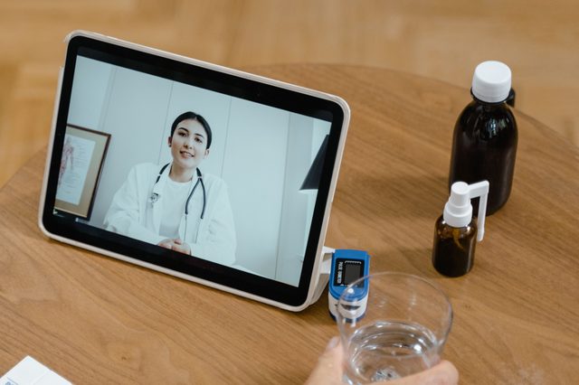 Una doctora aparece en la pantalla de una tableta, sobre una mesa con frascos de medicina, un oxímetro y un vaso de agua.